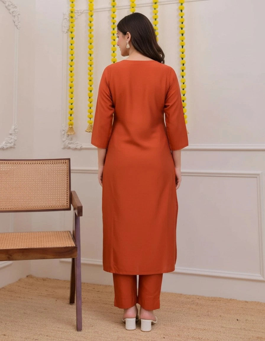 Woman wearing an orange kurta with pants standing in a room with a wooden bench and decorative wall.